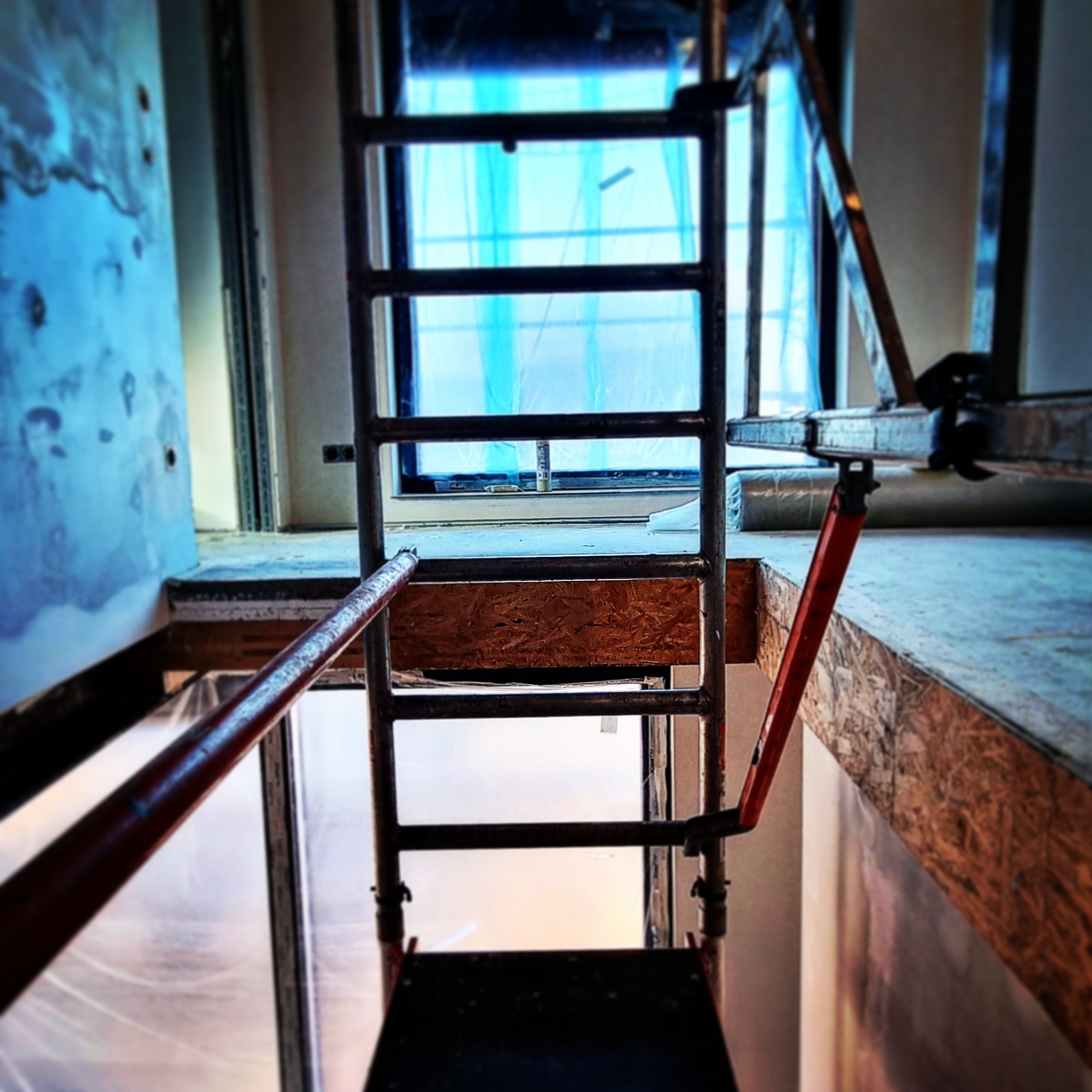 Wnętrze w trakcie prac wykończeniowych z widocznym rusztowaniem, zabezpieczonym oknem i fragmentem ściany z niedokończonymi pracami.