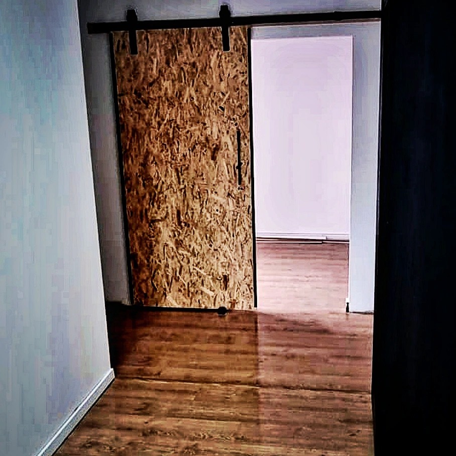 Przesuwne drzwi z płyty OSB na czarnej szynie, widoczne z boku w korytarzu z panelami podłogowymi, prowadzące do pomalowanego na biało pokoju.