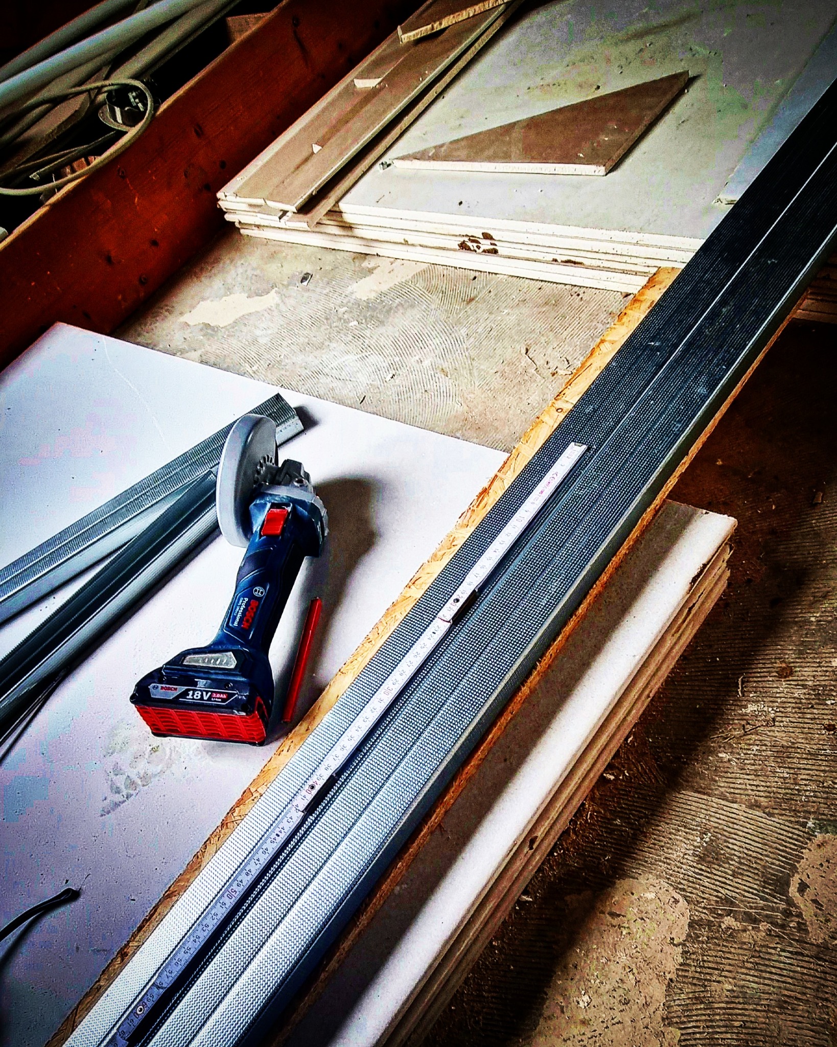 Narzędzia i materiały do montażu płyt kartonowo-gipsowych: szlifierka kątowa akumulatorowa Bosch, profil metalowy z miarką, płyta gipsowa, przygotowane do cięcia i montażu.