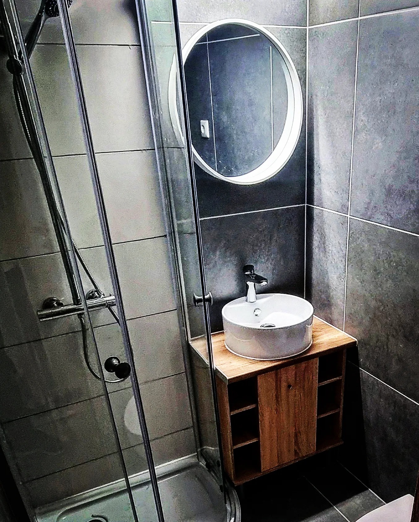 Nowoczesna łazienka z kabiną prysznicową, okrągłym lustrem z podświetleniem i umywalką nablatową na szafce z drewna w stylu loft, wykończona szarymi płytkami.