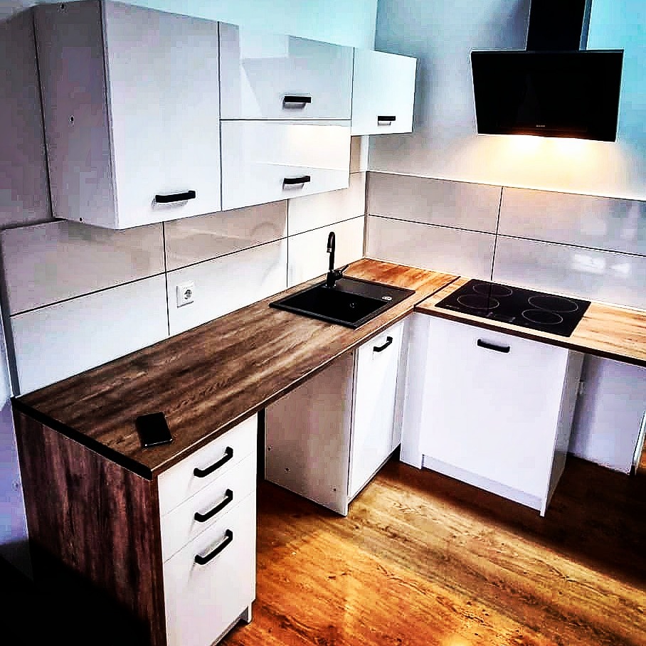 Nowoczesna kuchnia z białymi szafkami z czarnymi uchwytami, drewnianym blatem i czarnym zlewozmywakiem, płyta indukcyjna i okap, telefon leży na blacie.