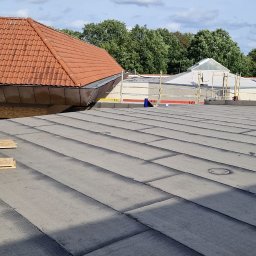 Zdzisław Głowacki - Widok z góry na płaski dach pokryty papą termozgrzewalną w trakcie prac dekarskich, z widocznym fragmentem dachu z czerwoną dachówką i rusztowaniami w tle.