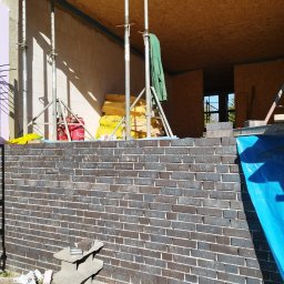 Zdzisław Głowacki - Wznoszenie ściany z ciemnej cegły klinkierowej, widoczne rusztowania, izolacja i materiały budowlane wewnątrz powstającego budynku.