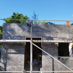 Zdzisław Głowacki - Wznoszenie ściany z ciemnej, licowej cegły na rusztowaniu, widoczne otwory okienne z drewnianymi ramami i robocza odzież zawieszona na rusztowaniu.