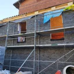 Budowa domu z ciemnej cegły klinkierowej, widoczne rusztowania, niezabudowane otwory okienne, częściowo zabezpieczone folią i płytą OSB, na pierwszym planie paleta z cegłą klinkierową Feldhaus...