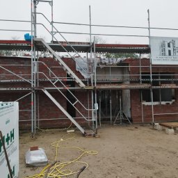 Zdzisław Głowacki - Budowa domu w stanie surowym otwartym, widoczne rusztowania, paleta cegieł, bloczki Ytong oraz baner firmy Haferkamp Gerüstbau.