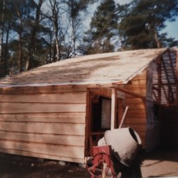 Zaklad usług budowlanych - Budowa małego, drewnianego domu w lesie. Widoczna betonowa mieszarka i konstrukcja dachu w trakcie budowy. Ściany z desek, surowy stan.