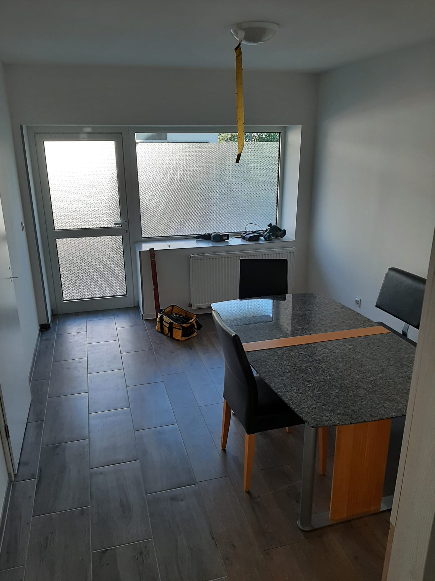 Wnętrze pokoju po remoncie z nową szarą podłogą z płytek imitujących drewno, stolikiem z ciemnym blatem i krzesłami, widoczna torba z narzędziami i poziomica przy drzwiach balkonowych.