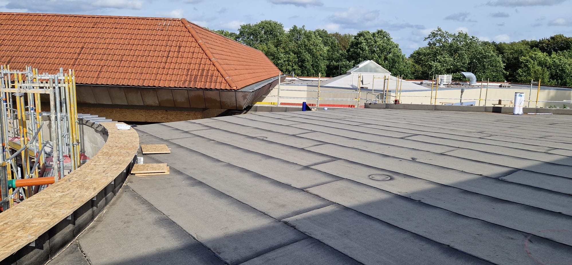 Widok z góry na płaski dach pokryty papą termozgrzewalną w trakcie prac dekarskich, z widocznym fragmentem dachu z czerwoną dachówką i rusztowaniami w tle.