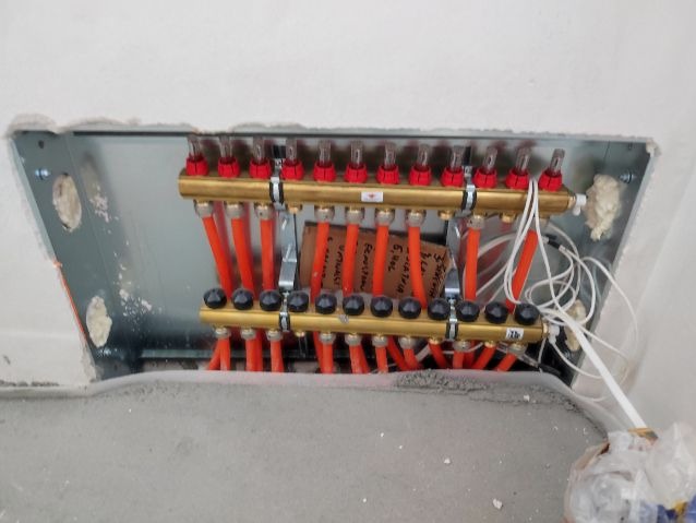 Rozdzielacz ogrzewania podłogowego z rurami w metalowej obudowie, widoczne zawory i przyłącza, częściowo zalany wylewką betonową, notatki na kartce.