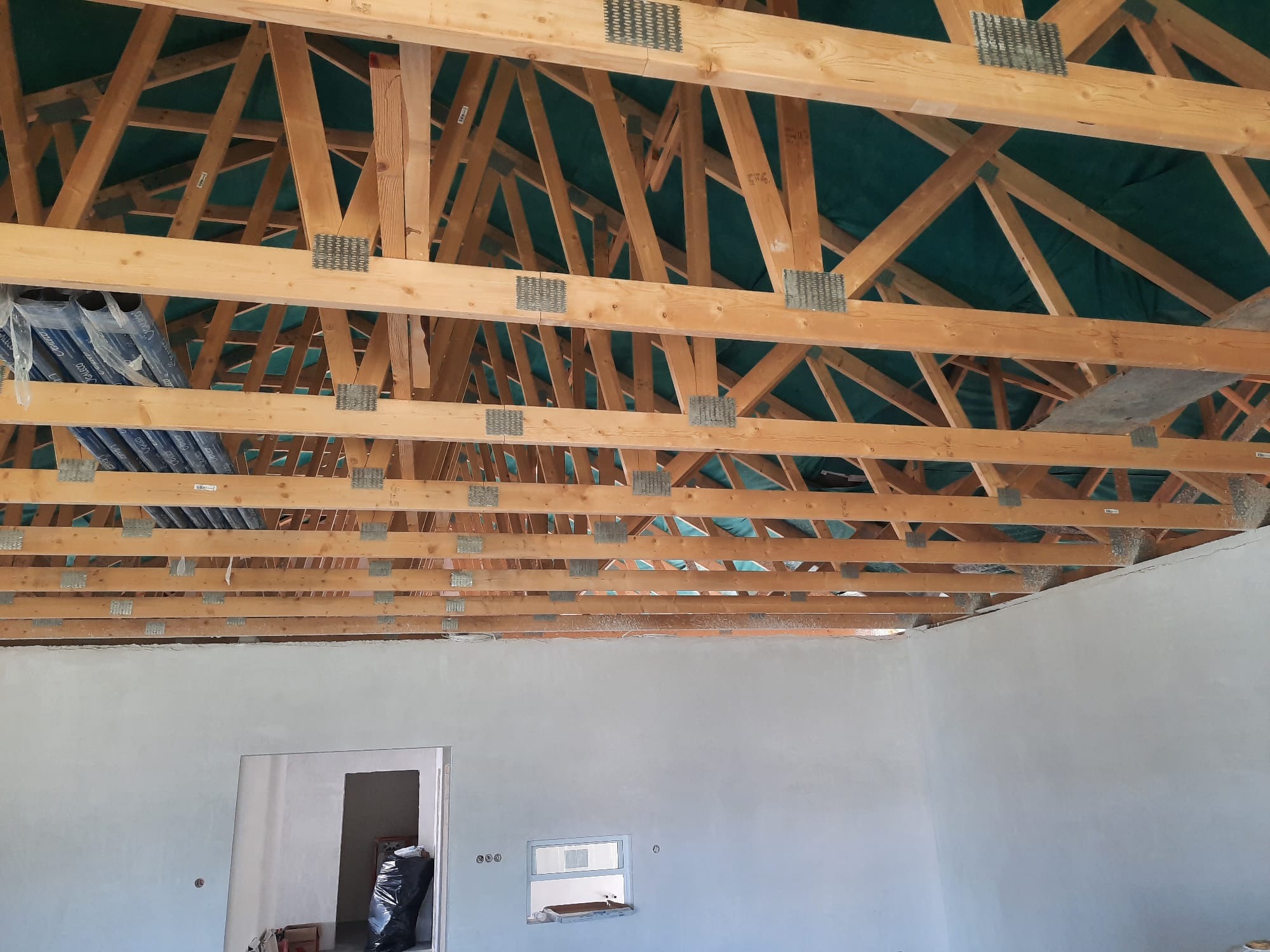 Wnętrze domu w trakcie budowy, widoczna drewniana konstrukcja dachu z metalowymi łącznikami oraz surowe, otynkowane ściany z otworem okiennym i drzwiowym.
