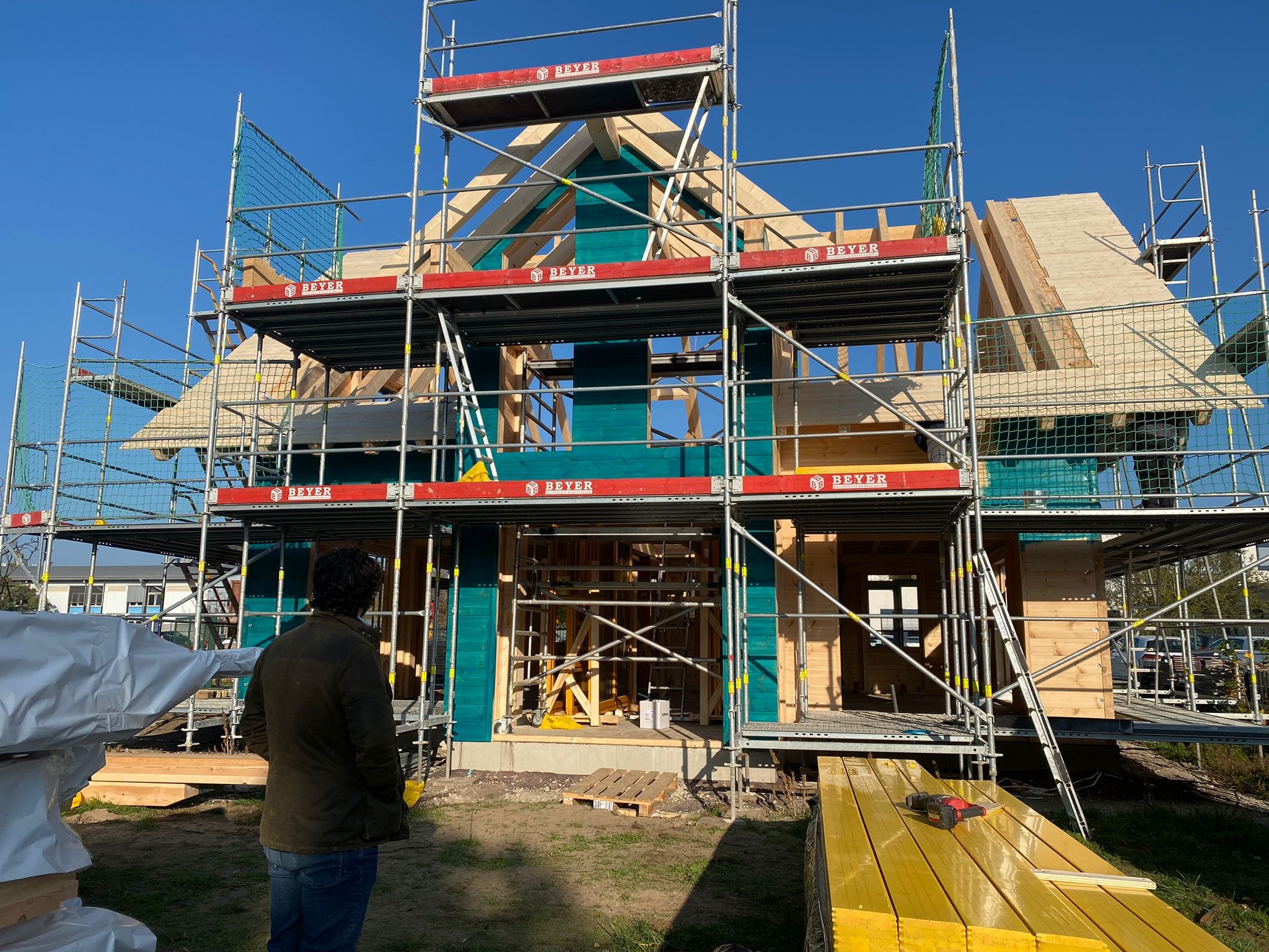 Wznoszenie konstrukcji drewnianej domu szkieletowego, widok od tyłu na mężczyznę obserwującego postęp prac, rusztowania otaczają budynek, na pierwszym planie stos żółtych desek i wkrętarka.