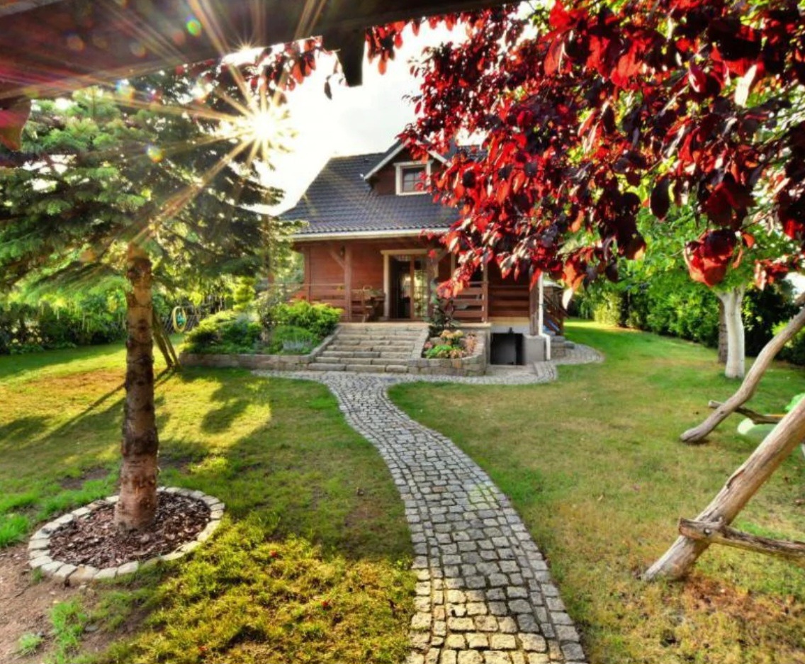 Urokliwy, parterowy dom z drewnianą elewacją i kamienną ścieżką w ogrodzie, oświetlony popołudniowym słońcem, z czerwonym drzewem po prawej stronie i zielonym drzewem po lewej stronie.