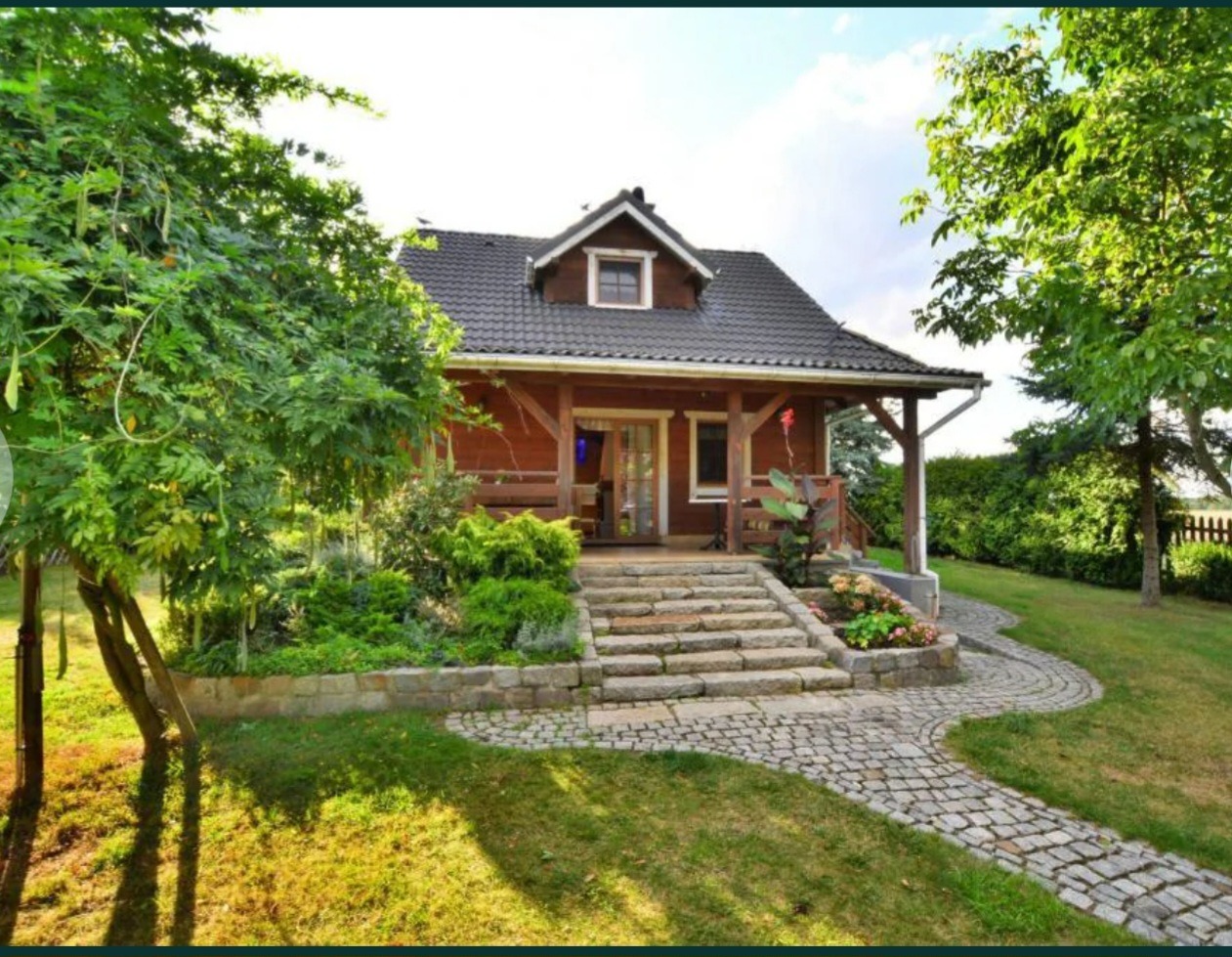 Drewniany dom z ciemnym dachem, kamienne schody i ścieżka z kostki brukowej, otoczony zielenią i drzewami w słoneczny dzień.
