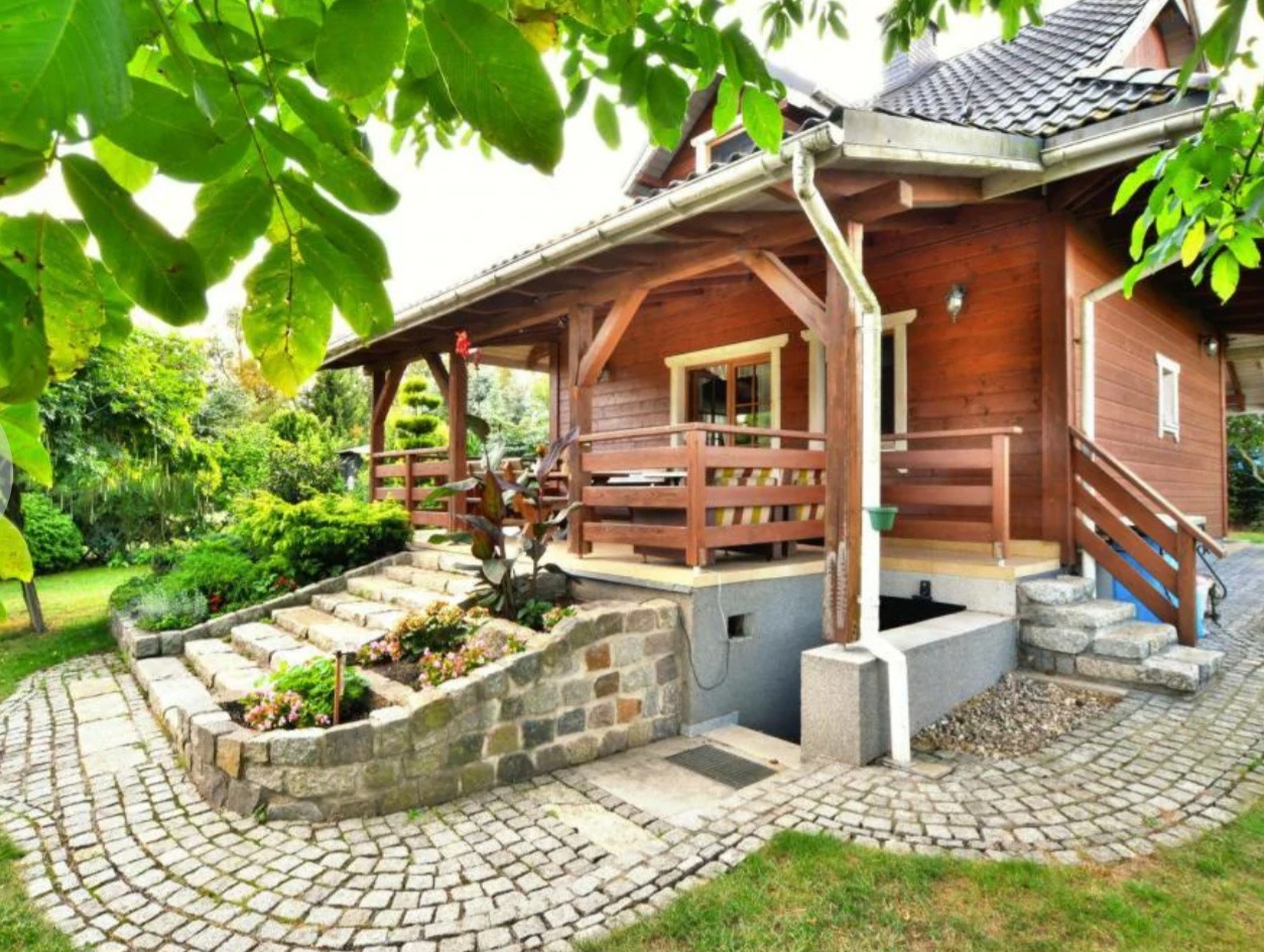 Drewniany dom z podpiwniczeniem i tarasem, kamienne schody z rabatami kwiatowymi oraz bruk wokół domu, widok z ogrodu.