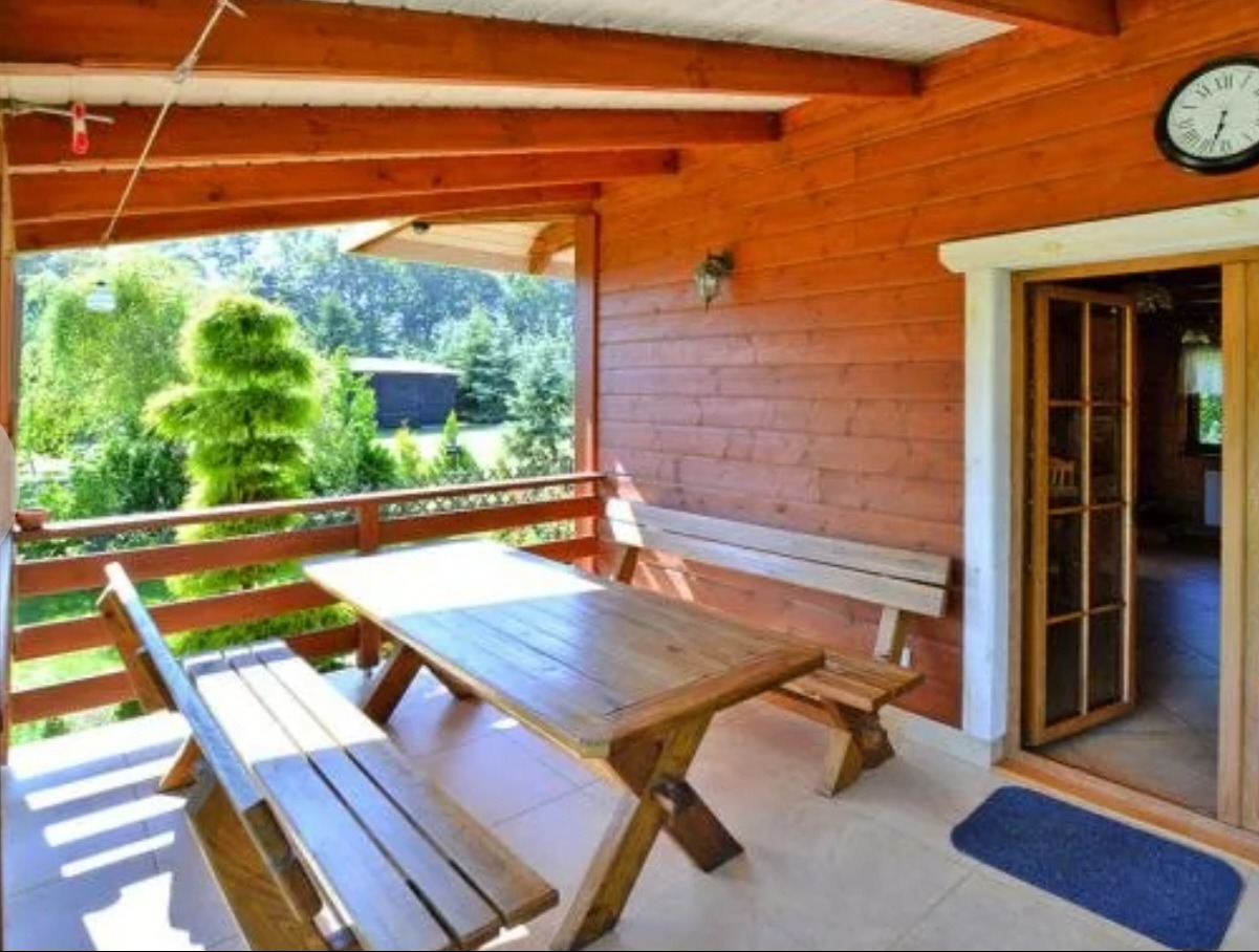 Zadaszony taras drewnianego domu z widokiem na ogród, wyposażony w stół i ławki z jasnego drewna, zegar na ścianie i niebieską wycieraczkę przed wejściem.