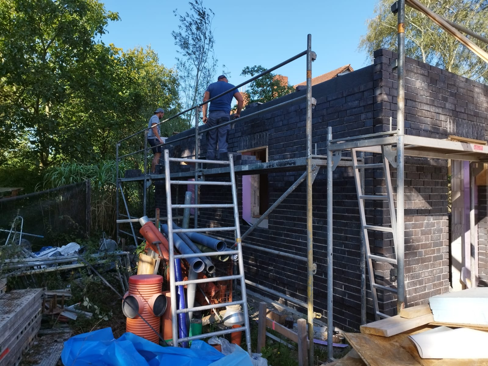 Dwóch murarzy na rusztowaniu w trakcie wznoszenia ścian z ciemnej cegły, widoczne materiały budowlane i elementy konstrukcyjne.