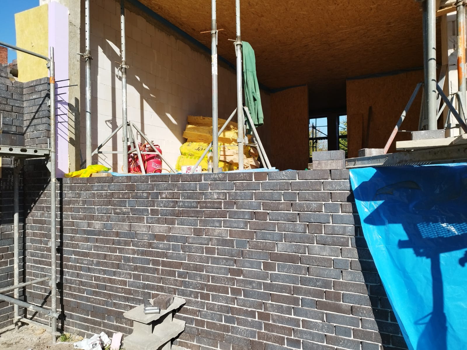 Wznoszenie ściany z ciemnej cegły klinkierowej, widoczne rusztowania, izolacja i materiały budowlane wewnątrz powstającego budynku.