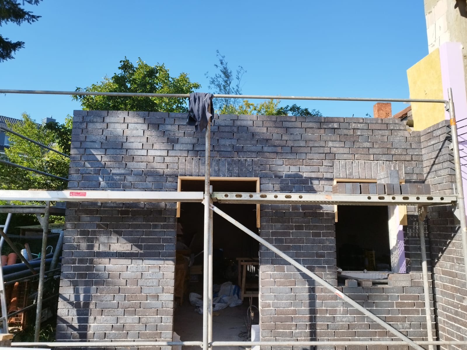 Wznoszenie ściany z ciemnej, licowej cegły na rusztowaniu, widoczne otwory okienne z drewnianymi ramami i robocza odzież zawieszona na rusztowaniu.