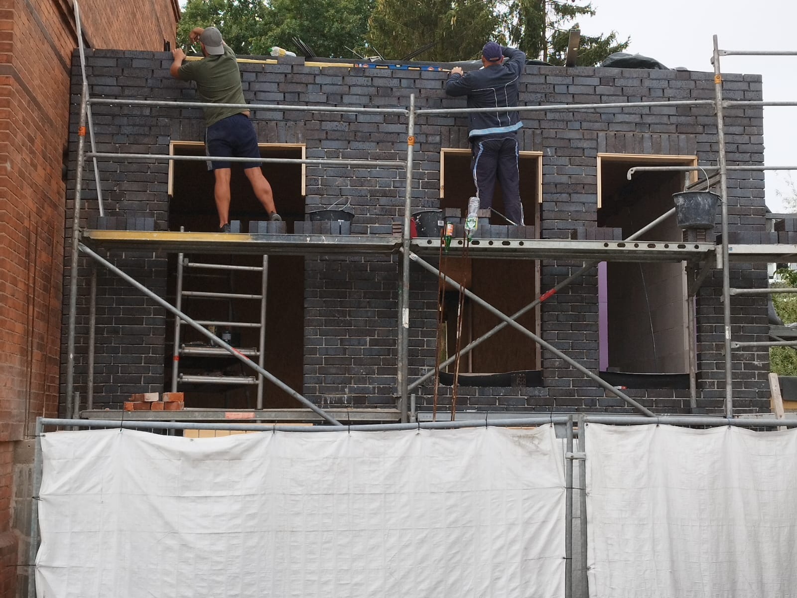 Dwóch murarzy pracuje na rusztowaniu przy wznoszeniu ściany z ciemnej cegły, widoczne otwory okienne, na dole biała plandeka.