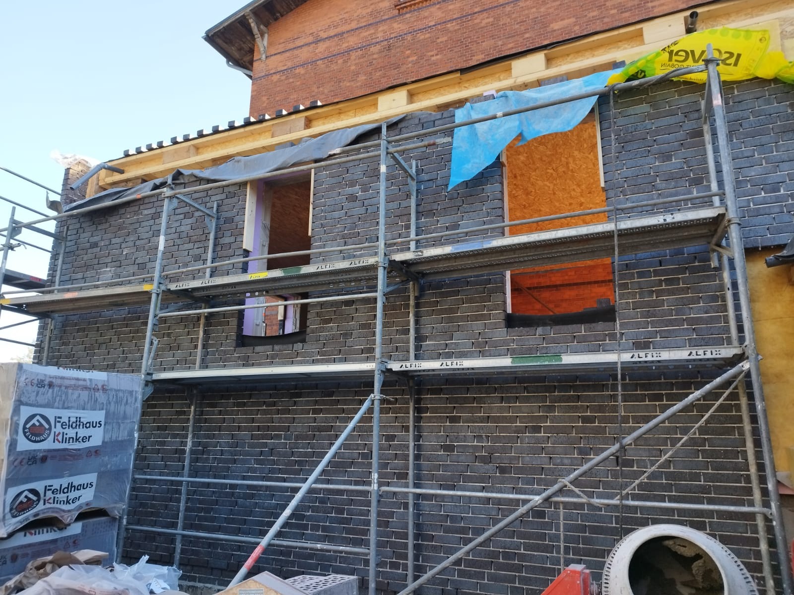 Budowa domu z ciemnej cegły klinkierowej, widoczne rusztowania, niezabudowane otwory okienne, częściowo zabezpieczone folią i płytą OSB, na pierwszym planie paleta z cegłą klinkierową Feldhaus...