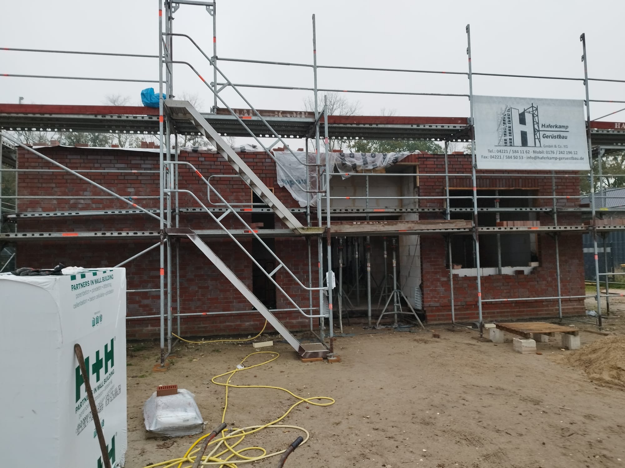 Budowa domu w stanie surowym otwartym, widoczne rusztowania, paleta cegieł, bloczki Ytong oraz baner firmy Haferkamp Gerüstbau.