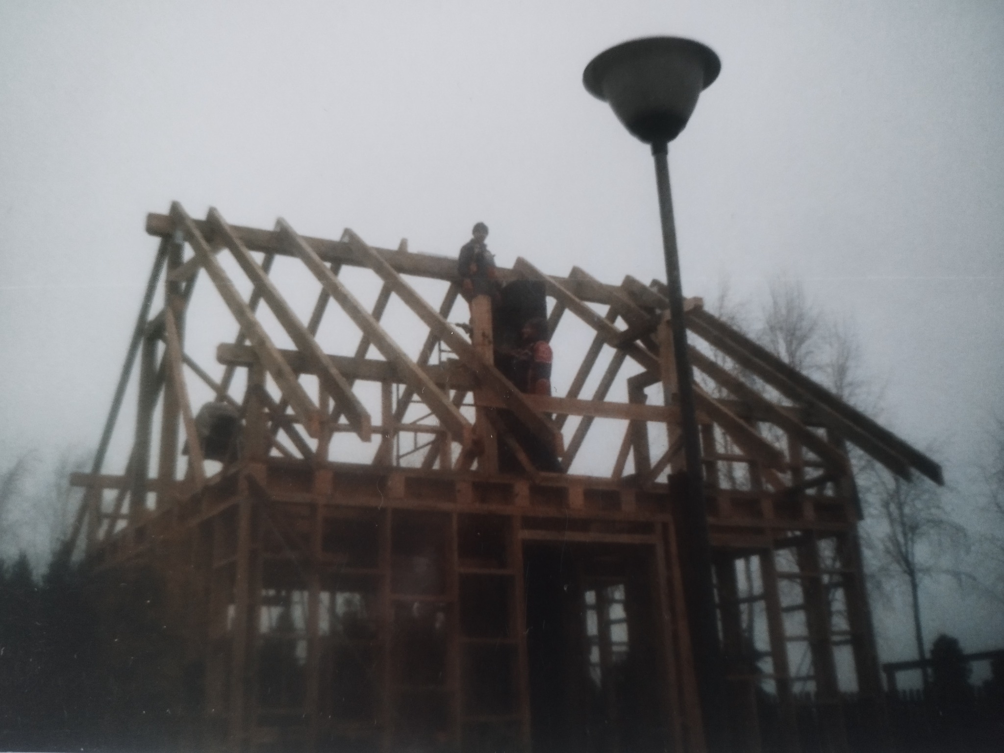 Wznoszenie drewnianej konstrukcji domu szkieletowego z widocznymi pracownikami na tle pochmurnego nieba i drzew. Ujęcie z dołu, widoczna latarnia uliczna.