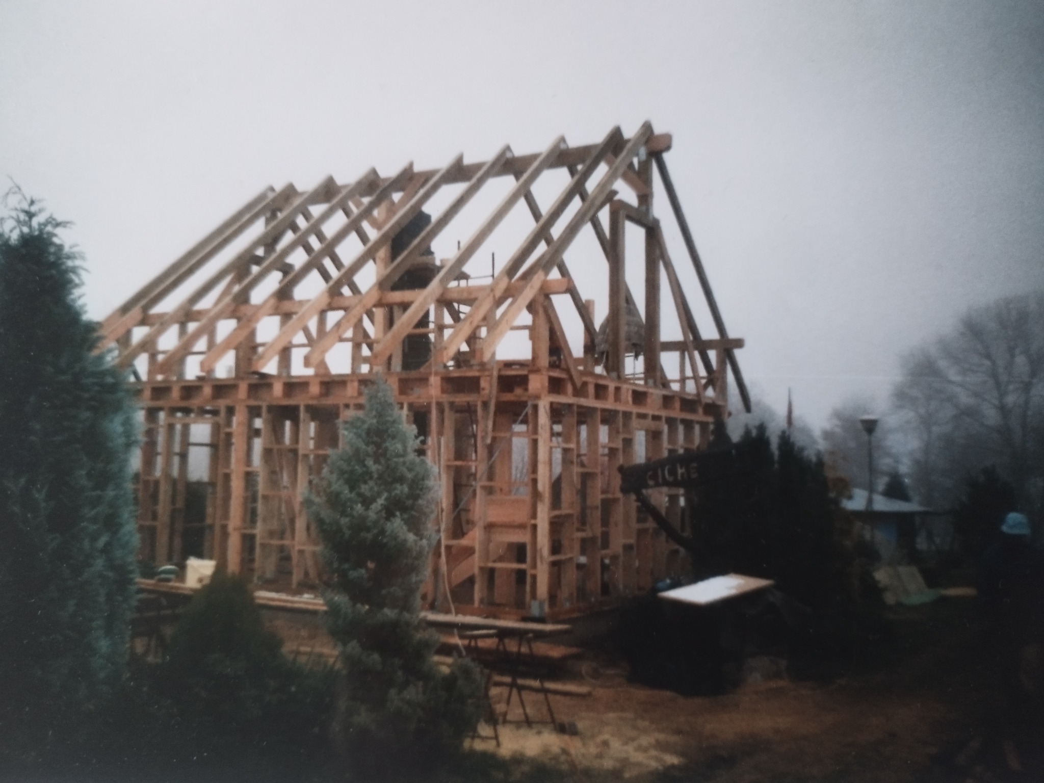 Konstrukcja drewniana domu w trakcie budowy, widoczne elementy szkieletu i dachu. W tle zieleń i pochmurne niebo. Na pierwszym planie drzewa iglaste.