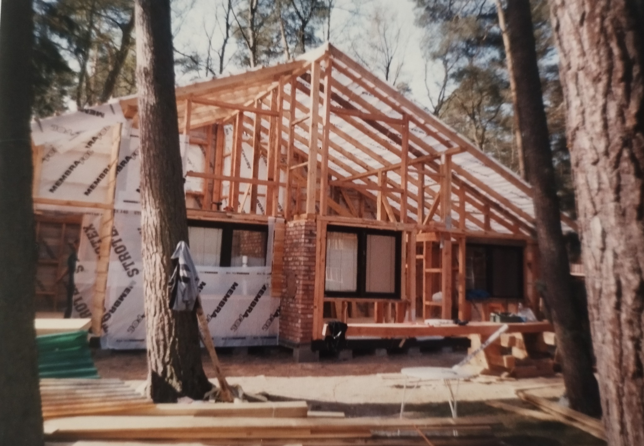 Budowa domu szkieletowego w lesie: drewniana konstrukcja dachu i ścian, folia ochronna, ceglany komin, okna z czarnymi ramami. Widok zza drzew.