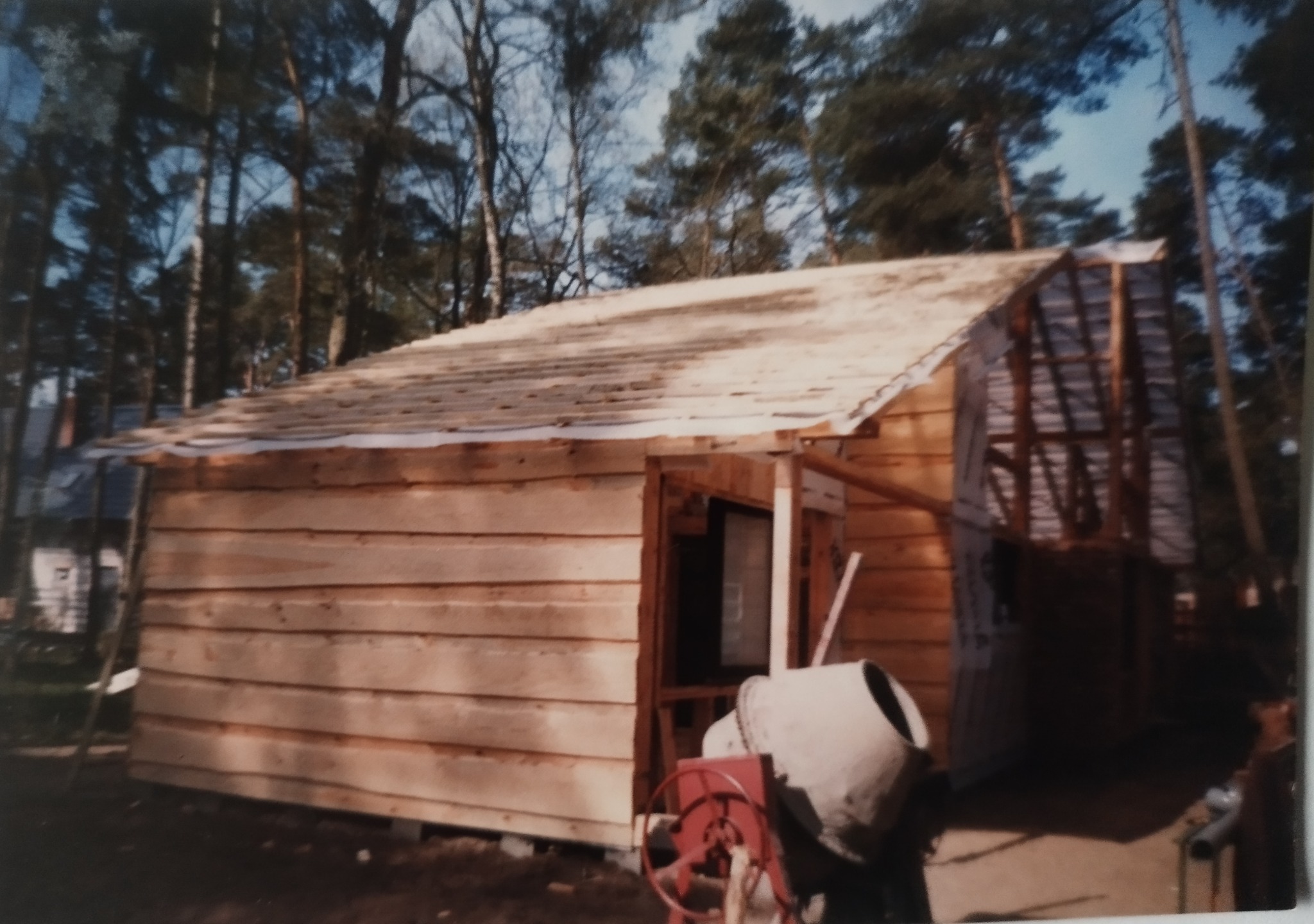 Budowa małego, drewnianego domu w lesie. Widoczna betonowa mieszarka i konstrukcja dachu w trakcie budowy. Ściany z desek, surowy stan.