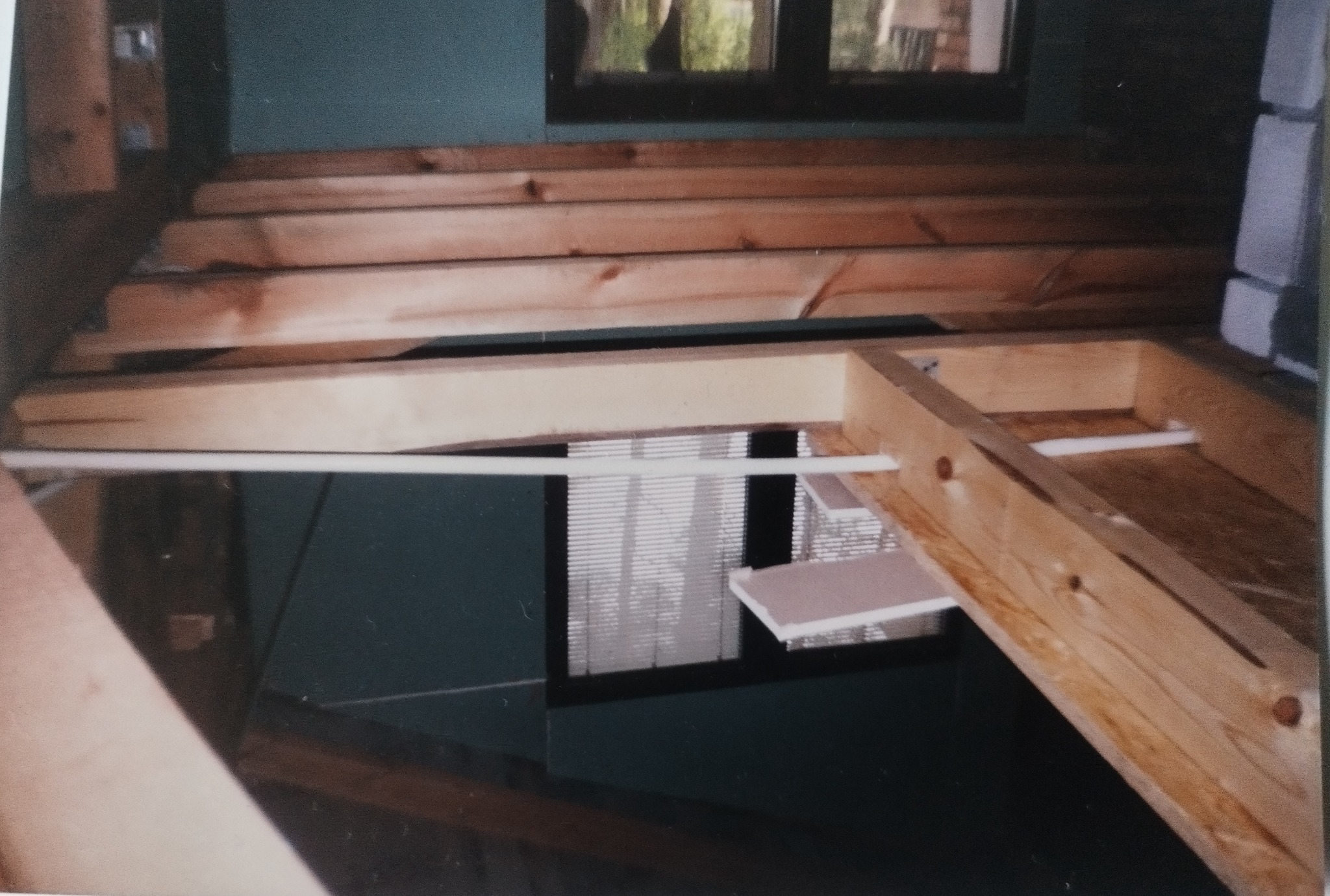 Wnętrze w trakcie budowy z drewnianą konstrukcją stropu i widocznymi elementami instalacji. Widok na okno z żaluzjami w tle. Surowy stan wykończenia.