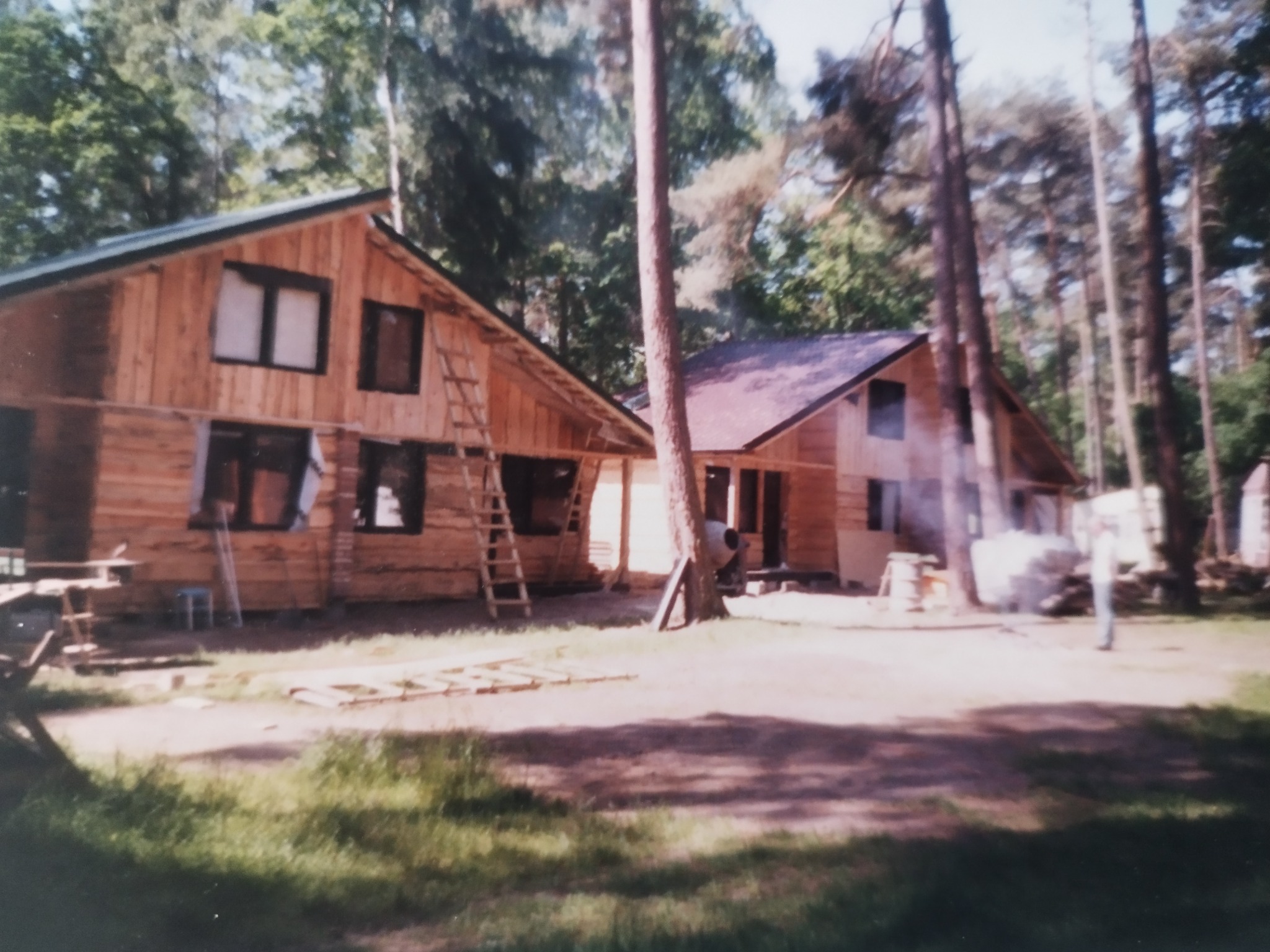 Dwa drewniane domy w trakcie budowy, z widocznymi rusztowaniami i drabinami, otoczone drzewami. Jeden dom z ciemnym dachem, drugi z jasnym. Prace budowlane w toku.