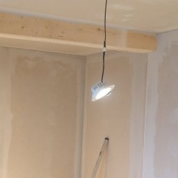 Mariusz Brzuszkiewicz - Ściana po szpachlowaniu, z widoczną drewnianą belką stropową i wiszącą lampą budowlaną. Widoczny fragment drabiny. Ściany przygotowane do dalszych prac wykończeniowych.