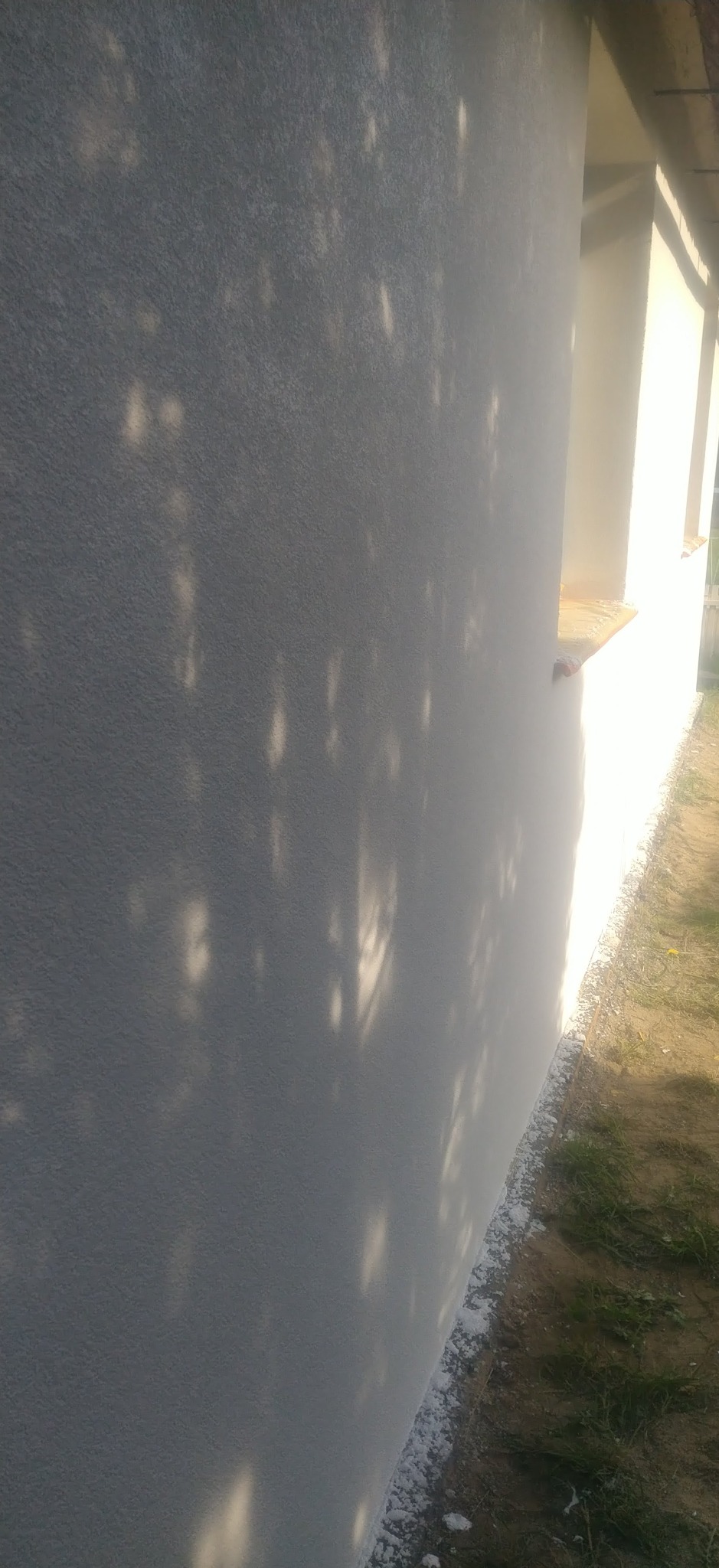 Świeżo pomalowana, biała elewacja budynku z widocznymi cieniami drzew, ukazana z boku w słoneczny dzień. Detal wykończenia zewnętrznego.