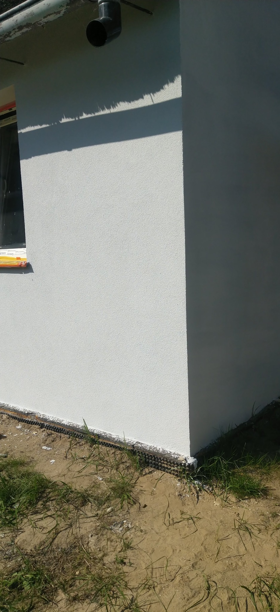 Świeżo pomalowana elewacja budynku w jasnym odcieniu, widoczny fragment okna i rynna, trawa przy fundamencie. Delikatna tekstura tynku.