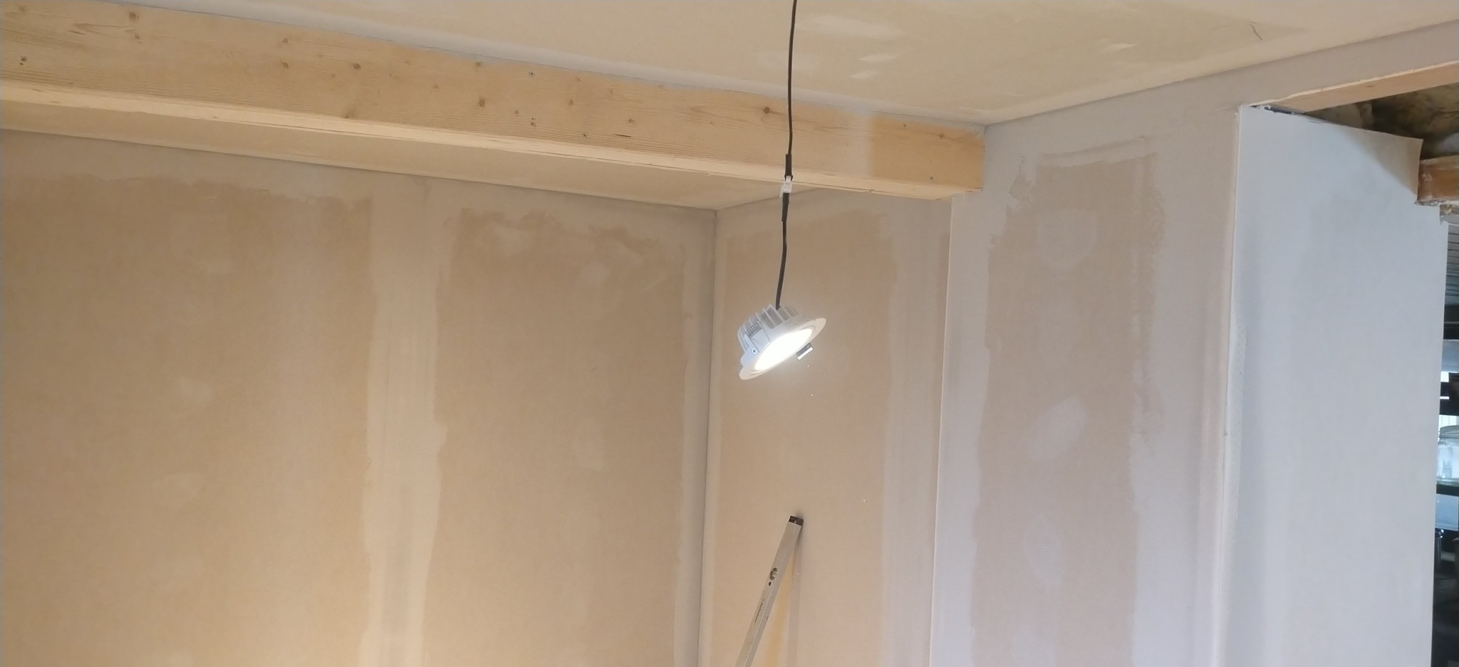 Ściana po szpachlowaniu, z widoczną drewnianą belką stropową i wiszącą lampą budowlaną. Widoczny fragment drabiny. Ściany przygotowane do dalszych prac wykończeniowych.