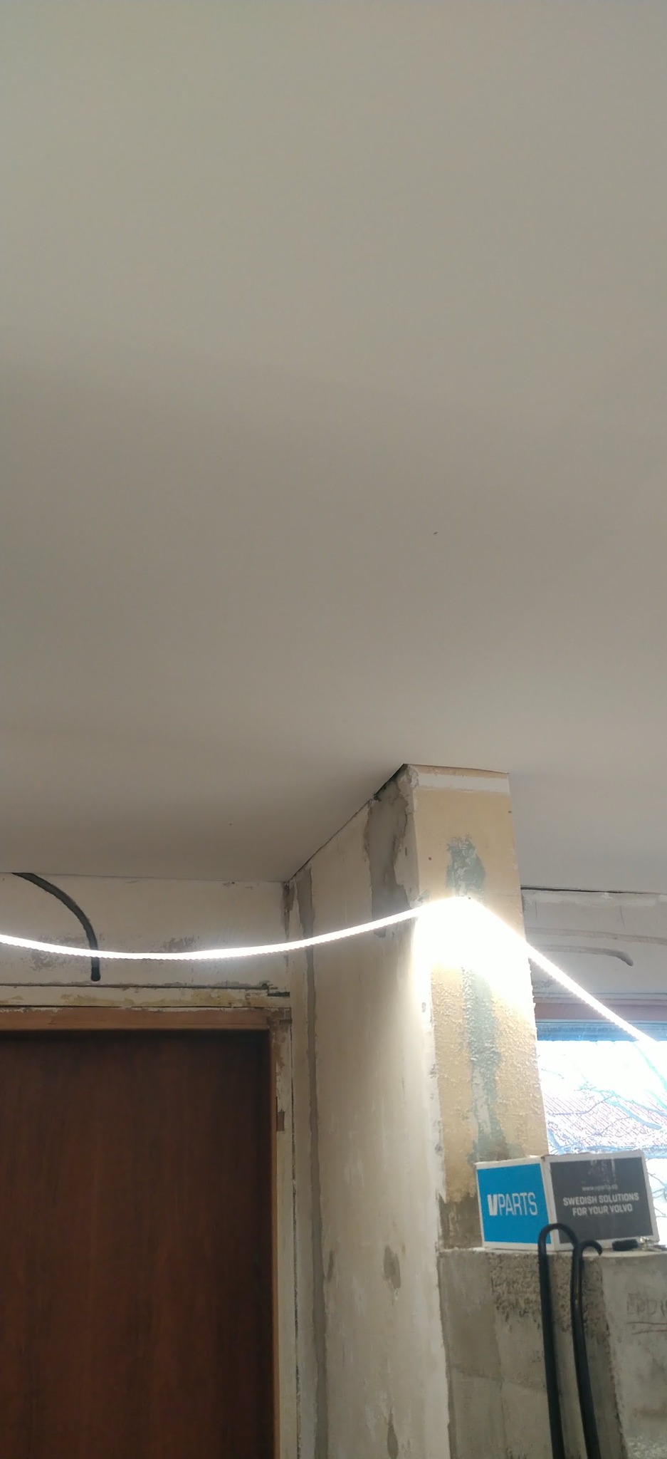 Narożnik ściany z widocznymi śladami po szpachlowaniu, oświetlony listwą LED. Fragment drzwi po lewej, okno z widokiem na zewnątrz po prawej.