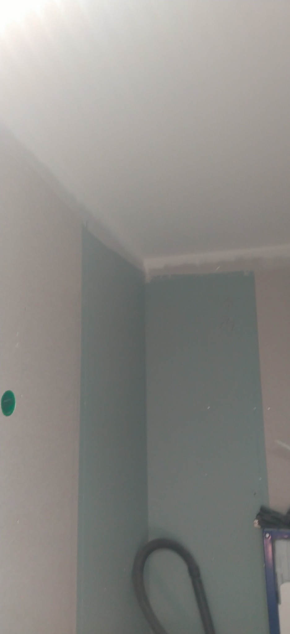 Narożnik pokoju z płytą kartonowo-gipsową, widoczne łączenia płyt na suficie i ścianach, przygotowane do dalszych prac wykończeniowych. Widoczny fragment odkurzacza.