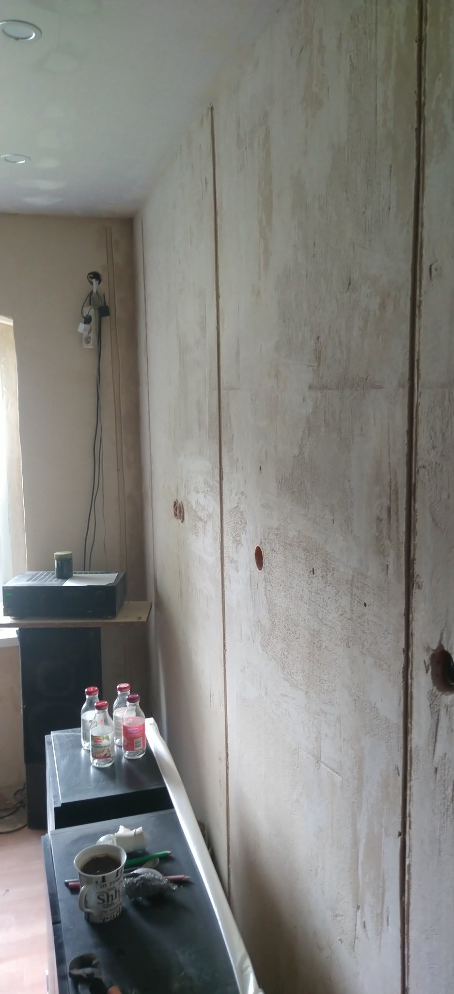 Ściana po szpachlowaniu, widoczne otwory na gniazdka elektryczne. Przy ścianie sprzęt audio i akcesoria malarskie. Prace wykończeniowe wnętrz w trakcie.