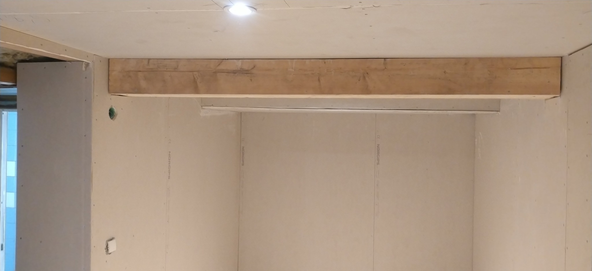 Wnętrze w trakcie prac wykończeniowych: ściany z płyt karton-gips, drewniana belka pod sufitem, widoczne otwory instalacyjne i fragment oświetlenia.