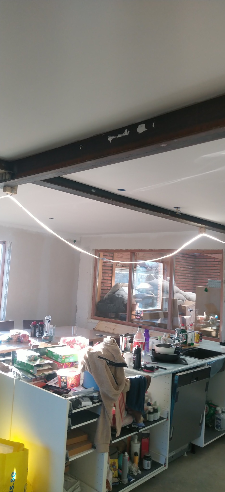 Wnętrze w trakcie prac wykończeniowych: widoczne belki stropowe, oświetlenie LED, ściany przygotowane do malowania i zagracona wyspa kuchenna.