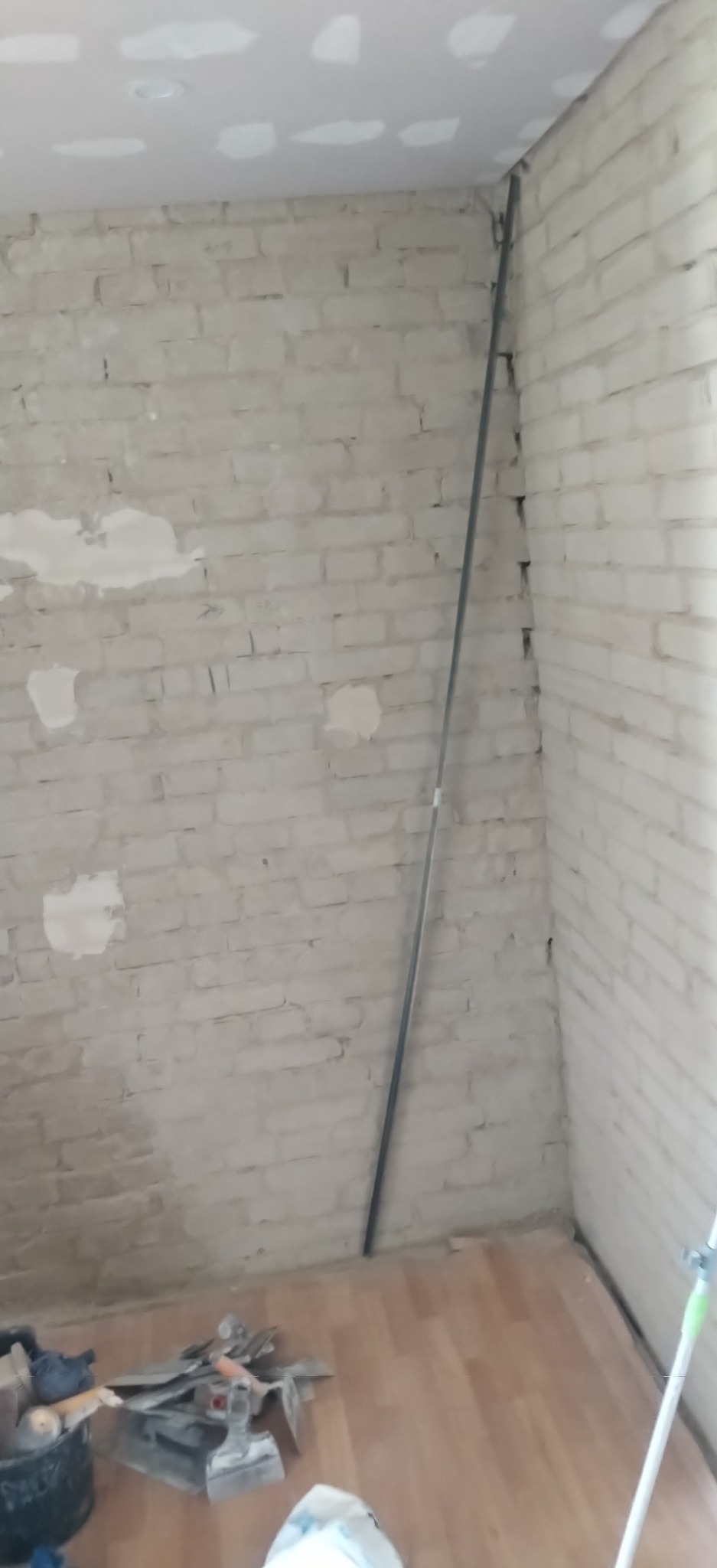Fragment wnętrza w trakcie prac wykończeniowych. Widoczna ceglana ściana z pozostałościami zaprawy, sufit z płyt gipsowych i narzędzia na podłodze.