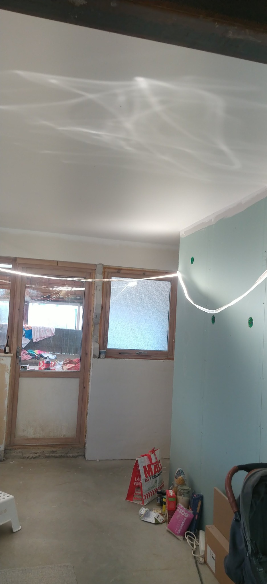 Wnętrze w trakcie remontu: ściana z płyt karton-gips, okna z drewnianą ramą, podłoga, ledy. Widoczne narzędzia i materiały budowlane. Całość w jasnej tonacji.