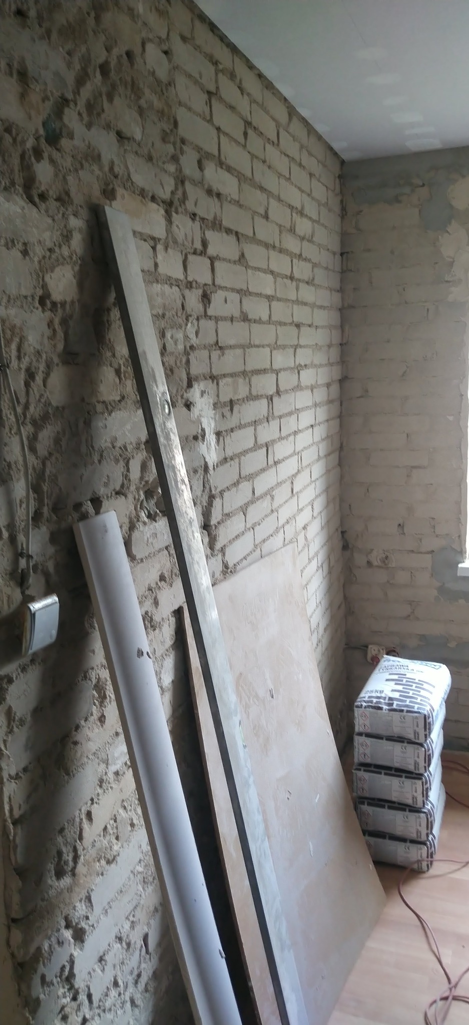Surowa ściana z cegły, przygotowana do prac wykończeniowych. Widoczne płyty karton-gips, listwa i worki z zaprawą. Fragment wnętrza w trakcie remontu.