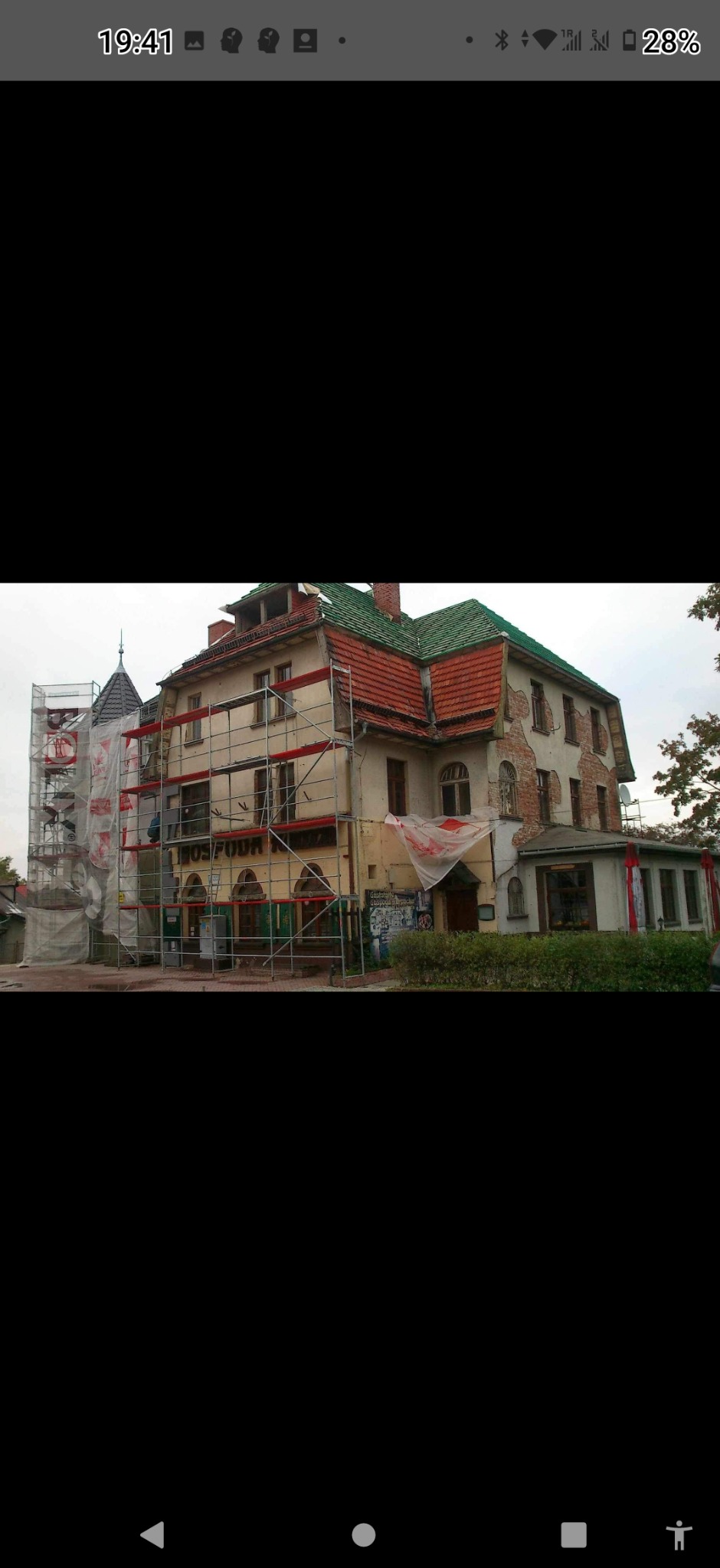 Renowacja elewacji starej kamienicy z zielonym dachem. Widoczne rusztowania, fragmenty odsłoniętej cegły i ślady graffiti. Częściowo zakryta folią.