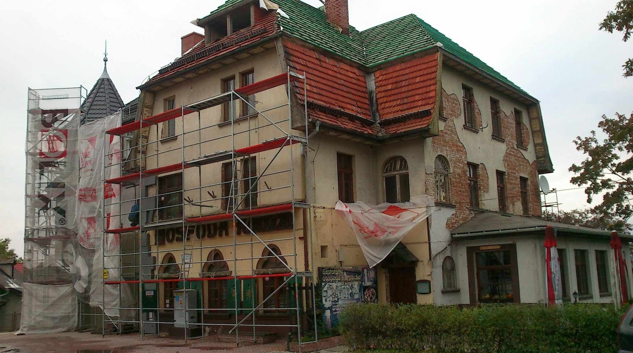 Budynek w trakcie renowacji z rusztowaniem i siatką ochronną. Widoczne fragmenty starej elewacji i dachu. Częściowo odsłonięta cegła.