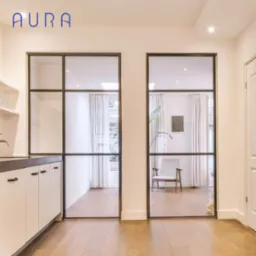 Nowoczesne, dwuskrzydłowe drzwi z czarną ramą i szklanymi panelami, oddzielające kuchnię od salonu, widok na wnętrze z drewnianą podłogą i minimalistycznym krzesłem.