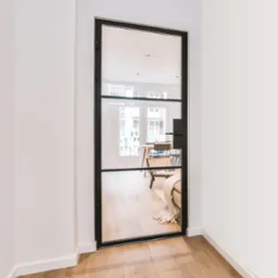 Czarne, stalowe drzwi z dużym szklanym panelem, widok na jasne wnętrze z drewnianą podłogą, krzesłem rattanowym i stołem w tle.