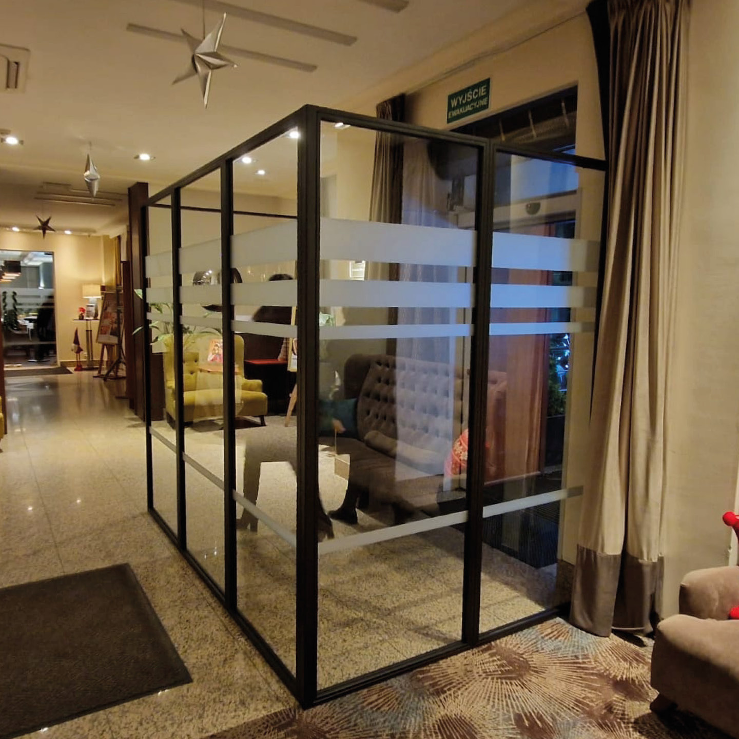 Szklana ścianka działowa z czarnymi profilami i poziomymi pasami matowej folii, oddzielająca przestrzeń w hotelowym wnętrzu z widocznymi sofami i wykładziną.