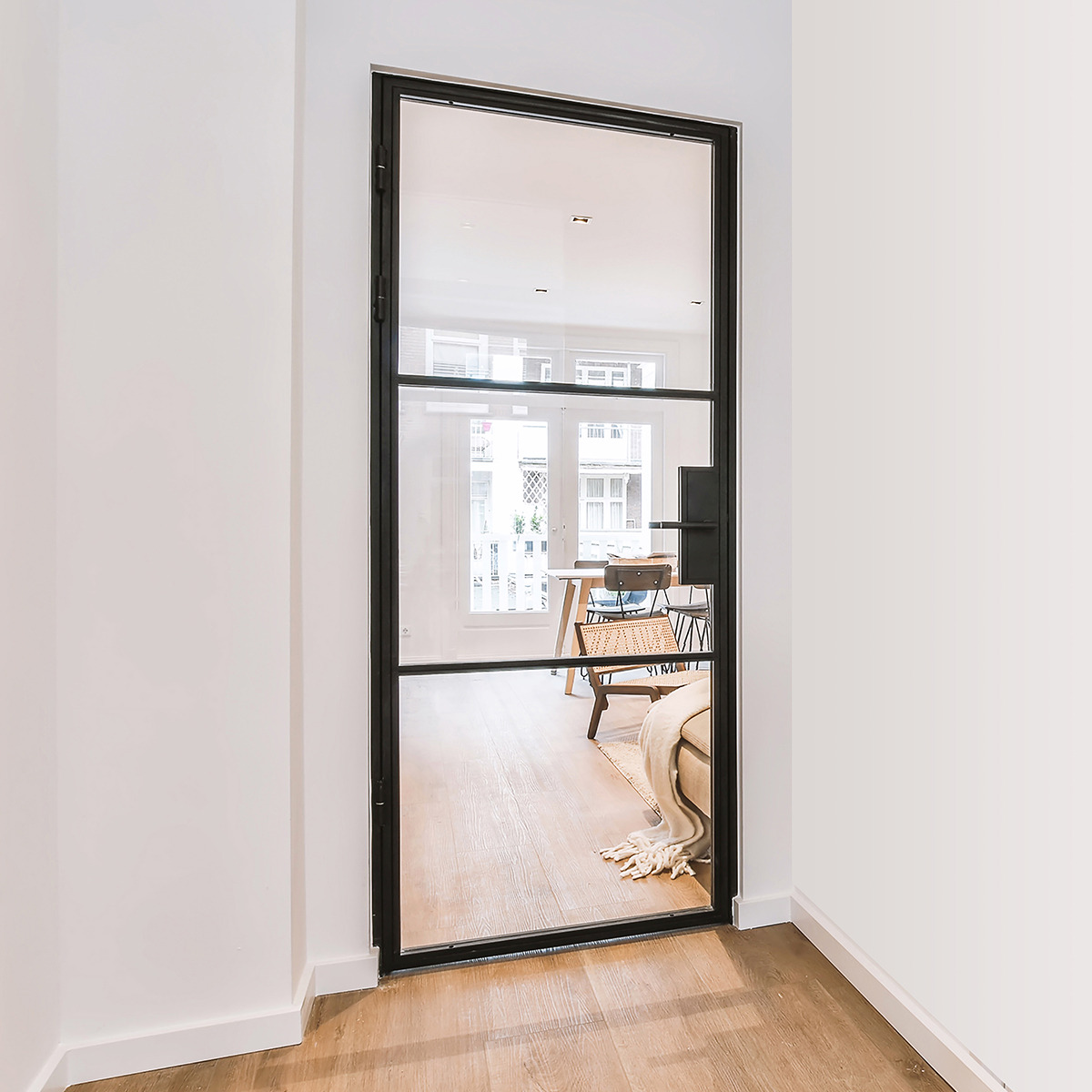 Czarne, stalowe drzwi z dużym szklanym panelem, widok na jasne wnętrze z drewnianą podłogą, krzesłem rattanowym i stołem w tle.